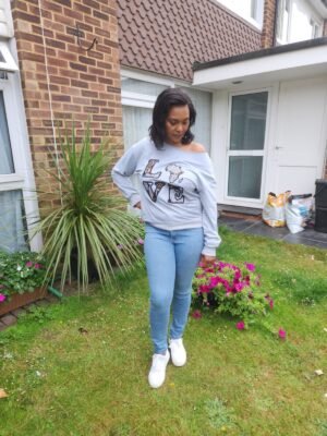 LOVE Africa Off-Shoulder Sweatshirt "Woman wearing a light grey off-the-shoulder sweatshirt with Ankara appliqué reading 'LOVE' and an Africa-shaped 'O', standing outdoors."