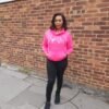 “LOVE Africa” Ankara Appliqué Hoodie – Hot Pink Woman wearing a hot pink hoodie with “LOVE” Ankara appliqué design, including the African continent in place of the "O", standing against a brick wall in casual black trousers and trainers.