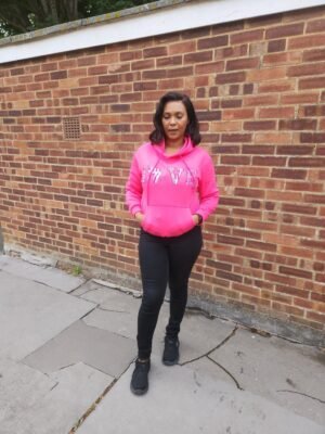 “LOVE Africa” Ankara Appliqué Hoodie – Hot Pink Woman wearing a hot pink hoodie with “LOVE” Ankara appliqué design, including the African continent in place of the "O", standing against a brick wall in casual black trousers and trainers.