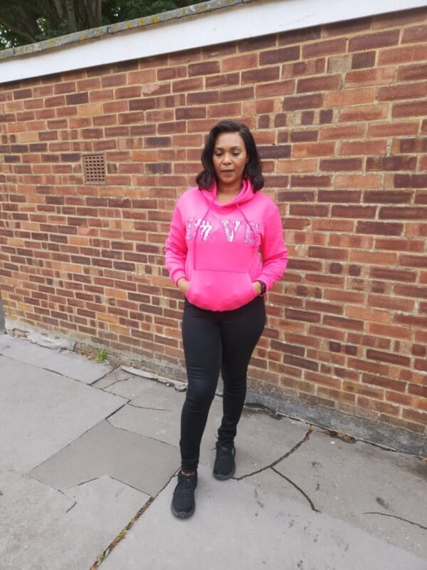 “LOVE Africa” Ankara Appliqué Hoodie – Hot Pink Woman wearing a hot pink hoodie with “LOVE” Ankara appliqué design, including the African continent in place of the "O", standing against a brick wall in casual black trousers and trainers.
