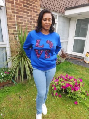Woman wearing a royal blue sweatshirt with pink and black Ankara leopard print appliqué spelling ‘LOVE’ with Africa-shaped ‘O’, standing in a garden.
