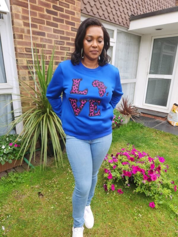 Ankara “LOVE Africa” Sweatshirt – Royal Blue | Leopard Print Appliqué Woman wearing a royal blue sweatshirt with pink and black Ankara leopard print appliqué spelling ‘LOVE’ with Africa-shaped ‘O’, standing in a garden.