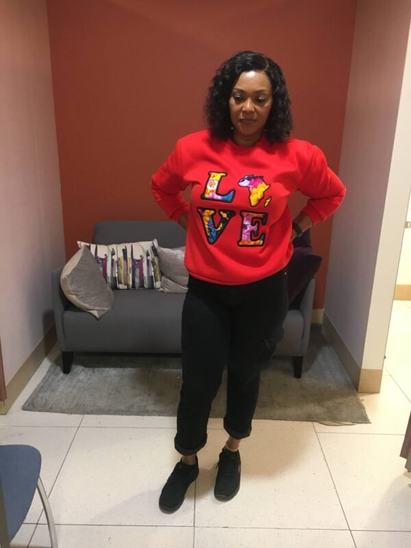 "LOVE Africa" Ankara Appliqué Sweatshirt – Red (Fleece-Lined) Woman wearing a bold red sweatshirt featuring 'LOVE' lettering in vibrant Ankara appliqué, with the letter 'O' creatively replaced by a stylized map of Africa. She is standing indoors with hands on hips, modeling the sweatshirt for product showcase. The design highlights African heritage through colorful, patterned cotton fabric stitched onto a soft polyester base."