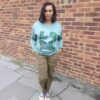 LOVE Africa Sweatshirt Sage Green Woman wearing sage green fleece-lined sweatshirt with Ankara “LOVE Africa” appliqué, standing in front of a brick wall.