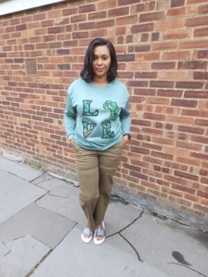 Woman wearing sage green fleece-lined sweatshirt with Ankara “LOVE Africa” appliqué, standing in front of a brick wall.